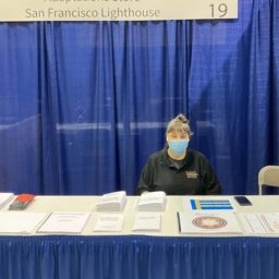 Adaptations store manager, Raqi Gomez, sits at the LightHouse table in the ACB exhibit hall. There are several Braille and tactile graphic products displayed on the table.