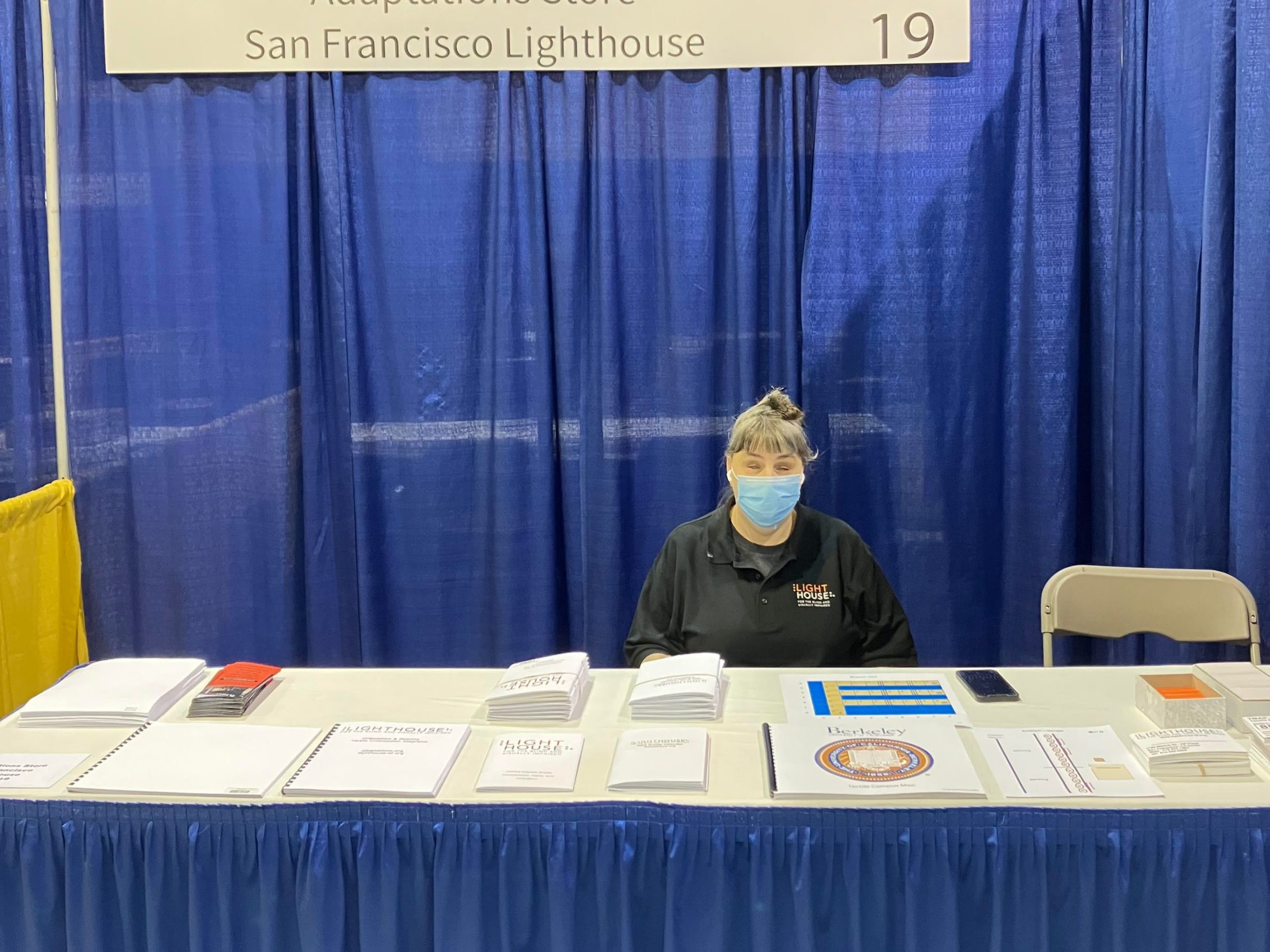 Adaptations store manager, Raqi Gomez, sits at the LightHouse table in the ACB exhibit hall. There are several Braille and tactile graphic products displayed on the table.