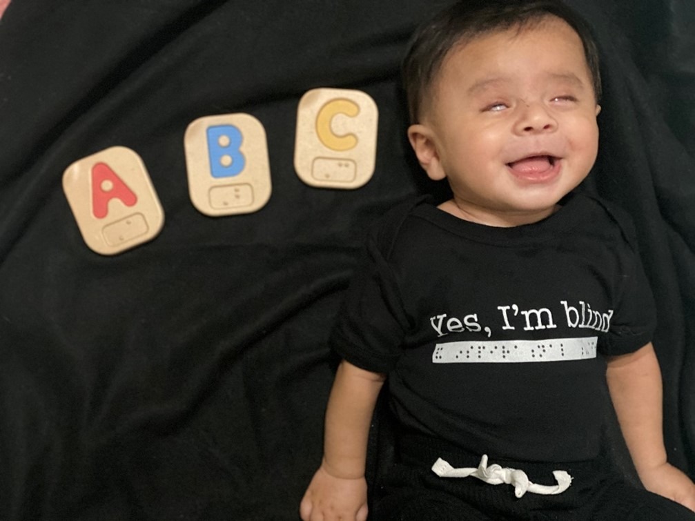  Baby boy is laying down next to ABC Braille blocks. He is wearing a onesie that says "Yes, I'm blind" in print and "and inspirational" in Braille dots