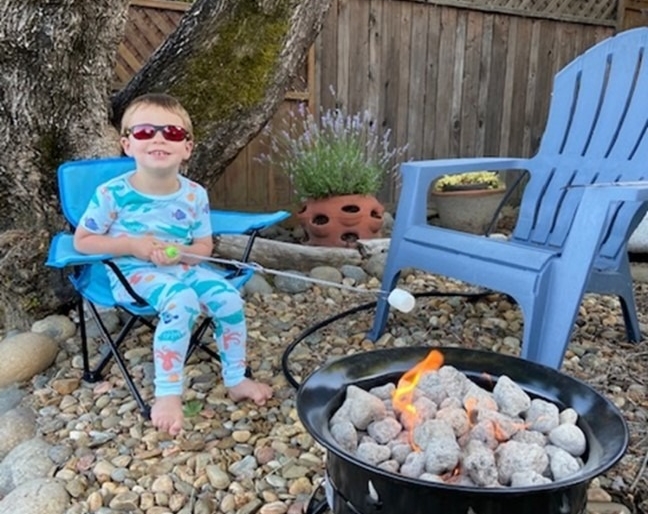 Little Learner sits on a chair in a backyard holding a marshmallow on a long stick over hot coals