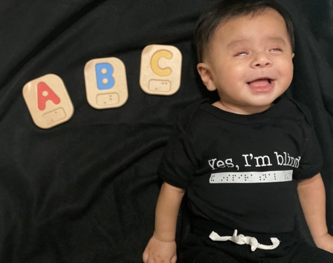 Little Learner is laying down next to ABC Braille blocks. His shirt reads, "Yes, I'm blind" in print and braille