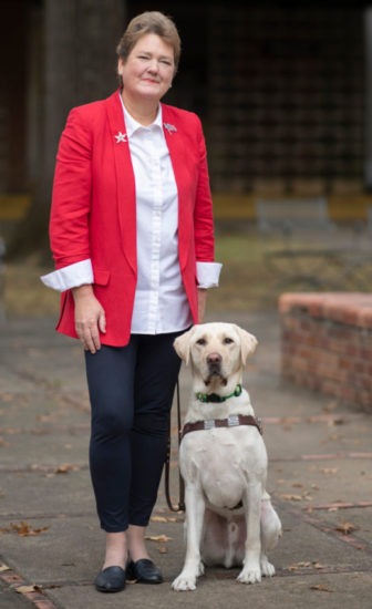 Sharon Giovinazzo in a dark pink suit with a white blouse: she stands with her guide dog, Pilot, a yellow lab
