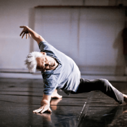 Jess Curtis is posed, mid-movement, with his right leg stretched out in front and his left leg stretched behind him. He is leaning back with his right hand on the floor and his left hand up in the air. His head is tilted sideways looking towards the camera