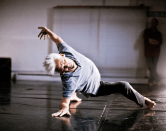 Jess Curtis is posed, mid-movement, with his right leg stretched out in front and his left leg stretched behind him. He is leaning back with his right hand on the floor and his left hand up in the air. His head is tilted sideways looking towards the camera