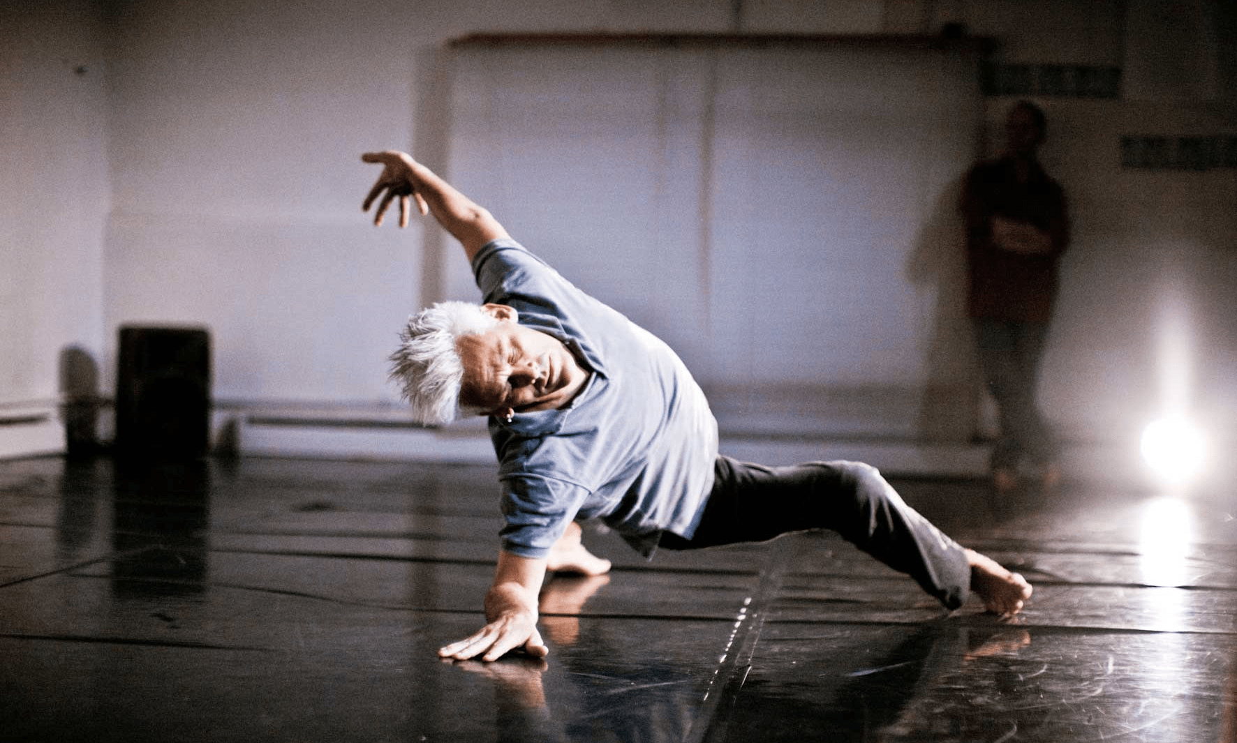 Jess Curtis is posed, mid-movement, with his right leg stretched out in front and his left leg stretched behind him. He is leaning back with his right hand on the floor and his left hand up in the air. His head is tilted sideways looking towards the camera