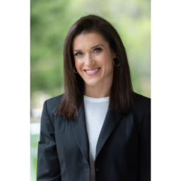 Rebecca Alexander is a woman with dark brown shoulder length hair and is wearing a white blouse under a black blazer.