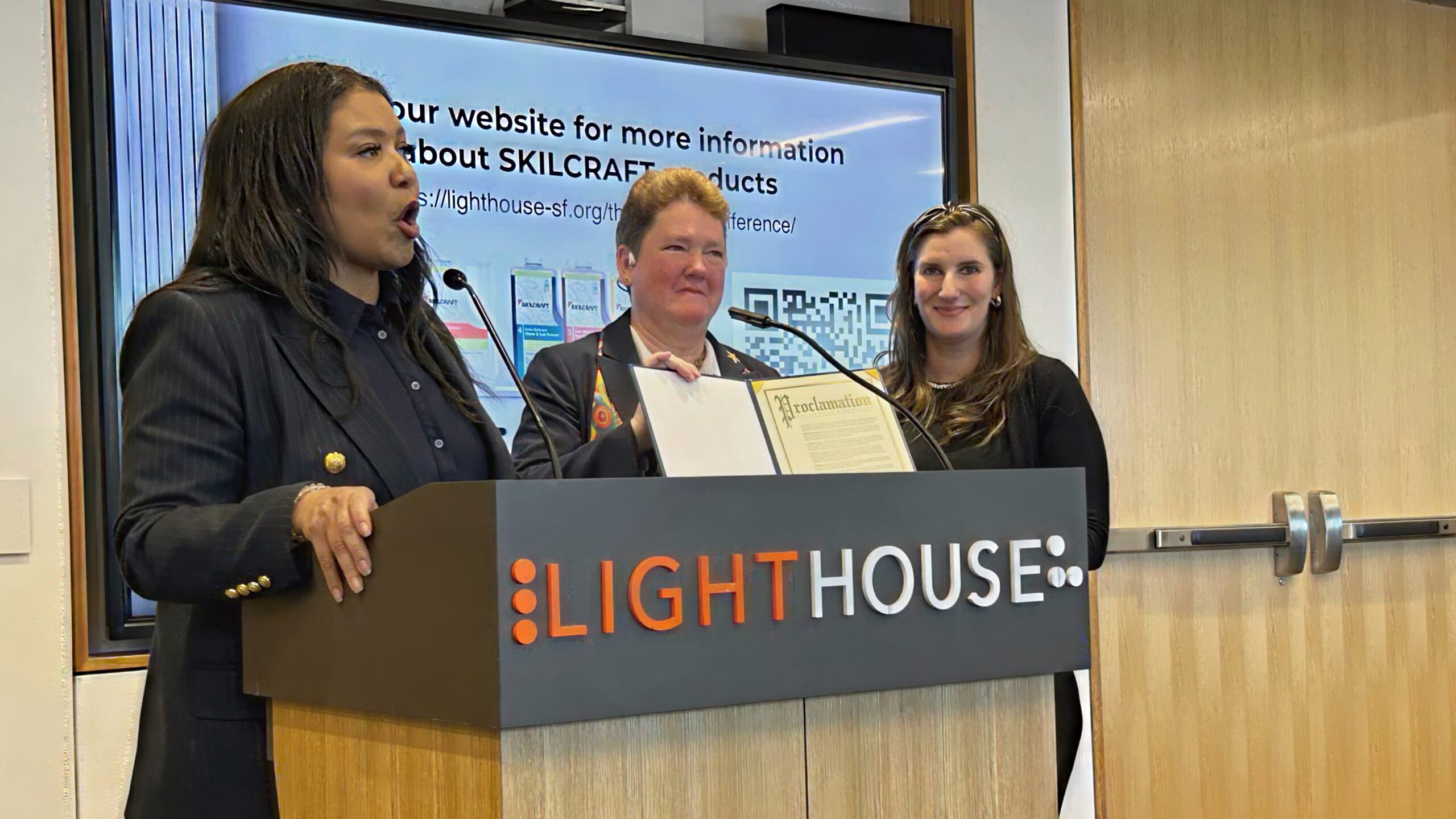 Mayor London Breed stands at a podium beside LightHouse CEO, Sharon Giovinazzo and Communications Specialist, Caitlin O’Malior