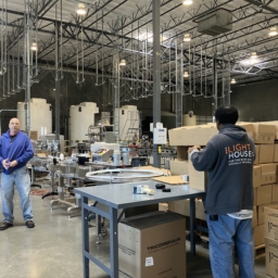 LightHouse Sirkin Center employee, Chris Peterson, stands on the manufacturing floor holding a bottled cleaning product while another employee wearing a LightHouse sweatshirt packages bottled products fresh off the manufacturing line for shipment.