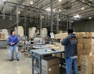 LightHouse Sirkin Center employee, Chris Peterson, stands on the manufacturing floor holding a bottled cleaning product while another employee wearing a LightHouse sweatshirt packages bottled products fresh off the manufacturing line for shipment.