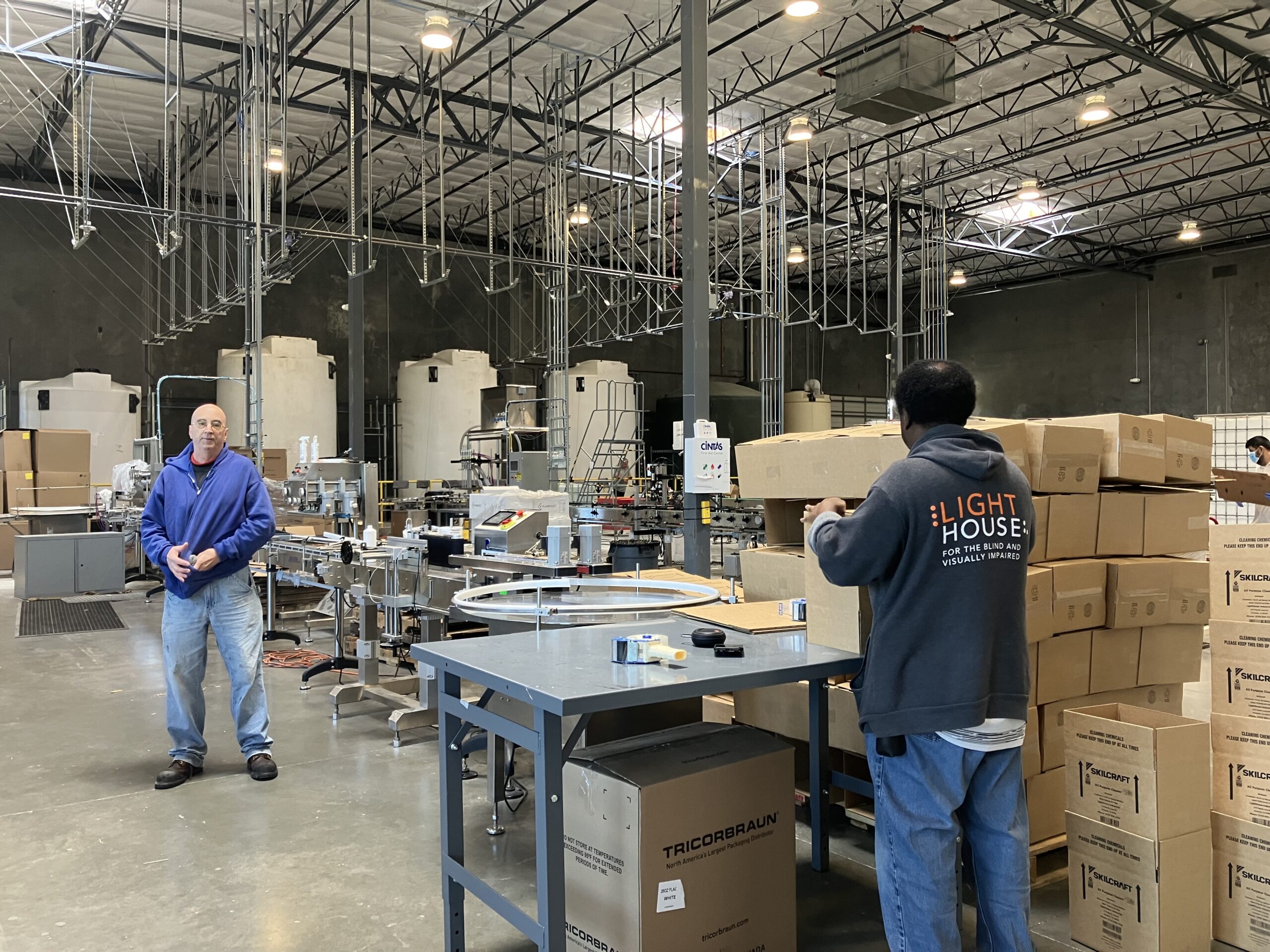 LightHouse Sirkin Center employee, Chris Peterson, stands on the manufacturing floor holding a bottled cleaning product while another employee wearing a LightHouse sweatshirt packages bottled products fresh off the manufacturing line for shipment.