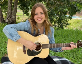 Meghan Downing sits outside on a black and white blanket and smiles while she strums a guitar.