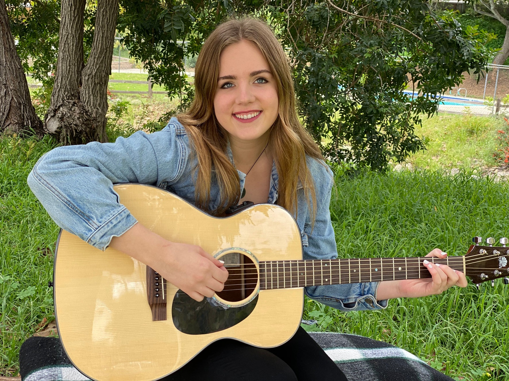 Meghan Downing sits outside on a black and white blanket and smiles while she strums a guitar.