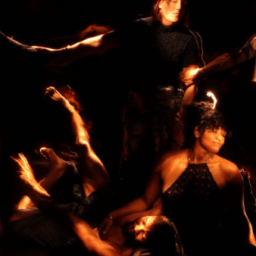 A photo from Into The Dark. Against a dark background, five figures appear, their bodies lit as if by a handheld flashlight. Three in the background link arms leaning away from each other as if in despair, or maybe rapture, while one figure sits on the ground in front, cradling the head of the fifth, prone figure, in her lap as the arm of the figure outstretched on the floor reaches up toward the trio standing behind
