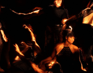 A photo from Into The Dark. Against a dark background, five figures appear, their bodies lit as if by a handheld flashlight. Three in the background link arms leaning away from each other as if in despair, or maybe rapture, while one figure sits on the ground in front, cradling the head of the fifth, prone figure, in her lap as the arm of the figure outstretched on the floor reaches up toward the trio standing behind