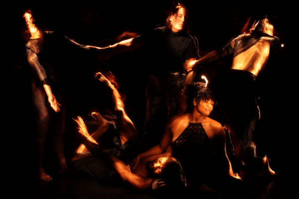 A photo from Into The Dark. Against a dark background, five figures appear, their bodies lit as if by a handheld flashlight. Three in the background link arms leaning away from each other as if in despair, or maybe rapture, while one figure sits on the ground in front, cradling the head of the fifth, prone figure, in her lap as the arm of the figure outstretched on the floor reaches up toward the trio standing behind