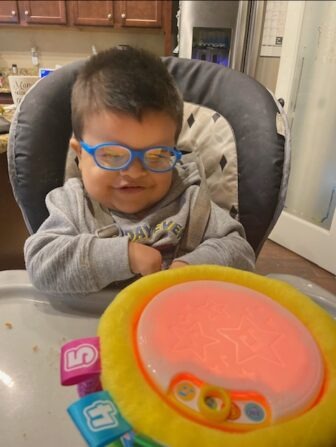 Little Learner Maximiliano, wearing glasses with blue frames, smiles. He sits behind a light-up toy.