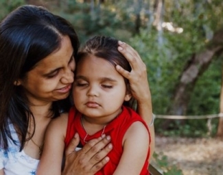 A mother holds her daughter with low vision in her lap. She cradles the little girl's head in one hand and holds her chest with the other to steady her
