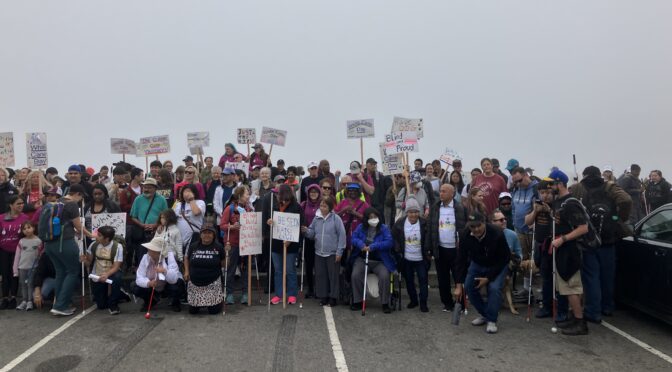LightHouse staff, students, volunteers and community members stand near the San Francisco side of the Golden Gate Bridge on White Cane Day in 2023. Many are holding signs with blind positive messages.
