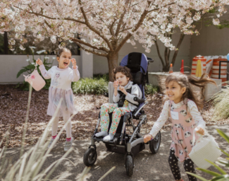 1. Three children smile holding Easter baskets, the child in the middle is a wheelchair user