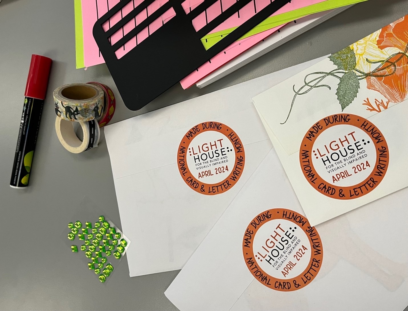 Art and adaptive letter writing supplies on a table with sealed envelopes and “Made During National Card & Letter Writing Month LightHouse April 2024” round stickers on the back.