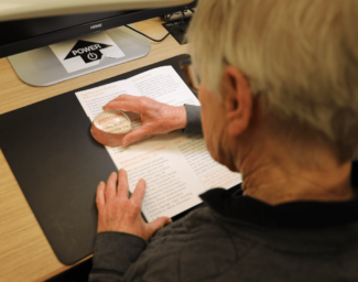 Seen from behind, a student sits at a desk and uses a dome magnifying glass to read text