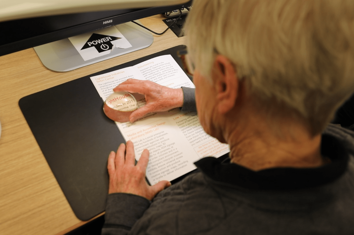 Seen from behind, a student sits at a desk and uses a dome magnifying glass to read text