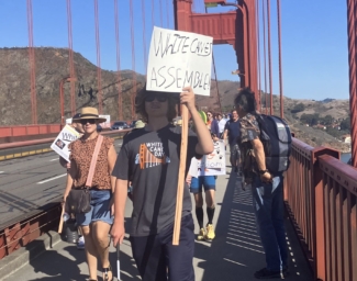White Cane Day marchers cross the Golden Gate Bridge. Center, a white cane user carries a sign that reads “White Canes Assemble”.