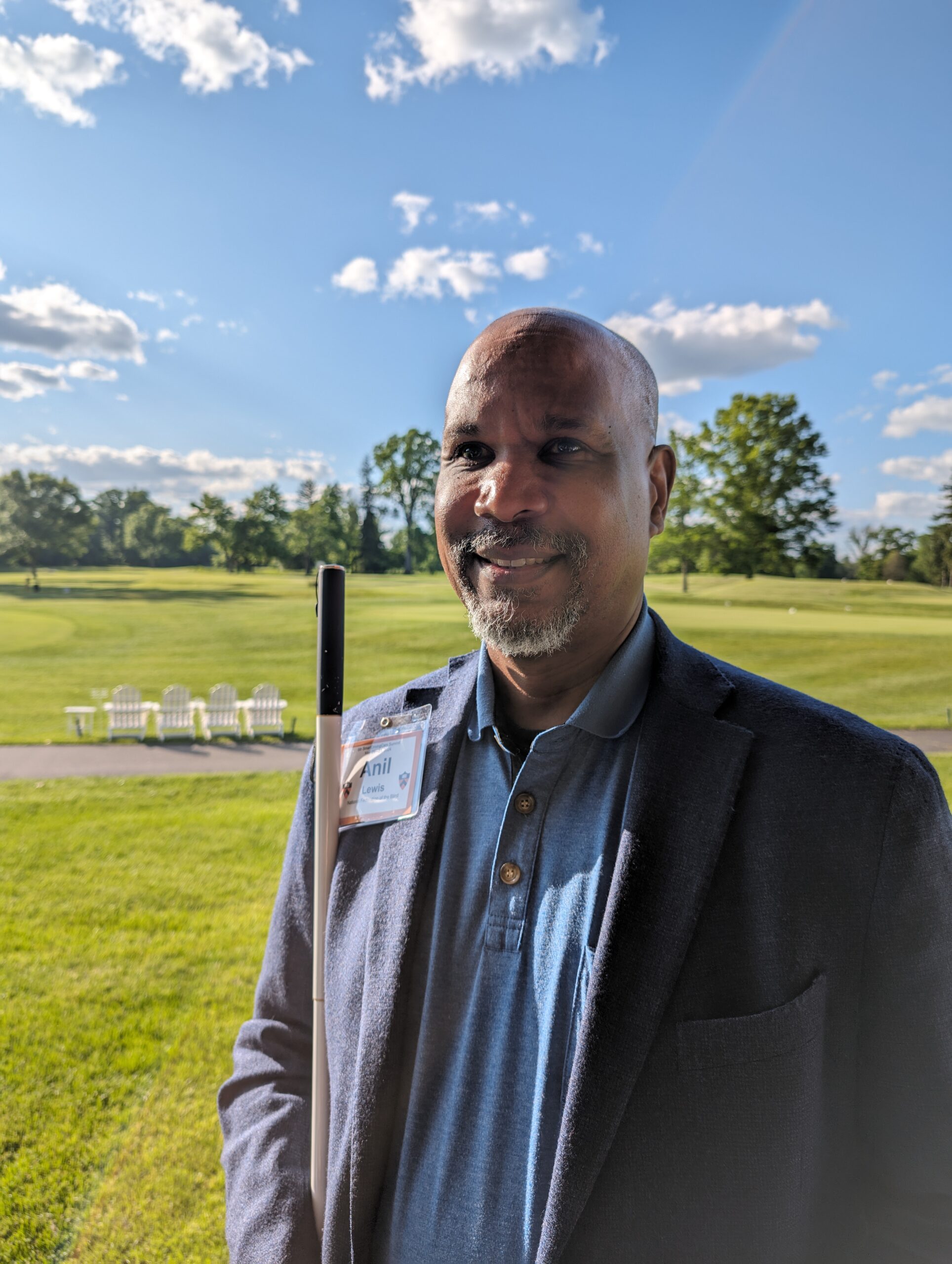 Anil Lewis stands outdoors in a field holding a long white cane on a partially cloudy day. Trees are seen in the distance.