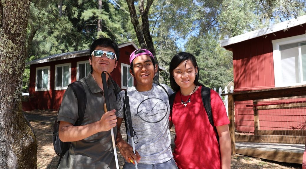 EHC-campers-1038x576 Three teens stand together in front of the lakeside cabins at Enchanted Hills Camp