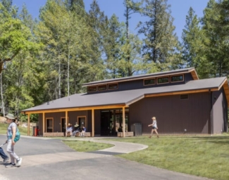 Two campers with white canes cross a cane-detectible path with the Blindness Training Center in the background. The gabled structure, situated with the forest beyond, welcomes campers through an expansive awning with campers chatting on large timber benches underneath. The dark metal-clad structure with large timber columns and redwood eaves evokes the materiality of the charred redwood bark of the trees that survived the fire.