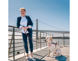 LightHouse CEO Sharon Giovinazzo stands with guide dog Pilot, a yellow lab, at the San Francisco Ferry terminal. A view of the San Francisco Bay Bridge can be seen behind her. She wears a blue pantsuit, a white-collared shirt and white heels. She holds Pilot’s leash while Pilot sits near her wearing a kerchief around the neck. The sun reflects off the top of Sharon’s short red hair as she smiles, mid-laugh.