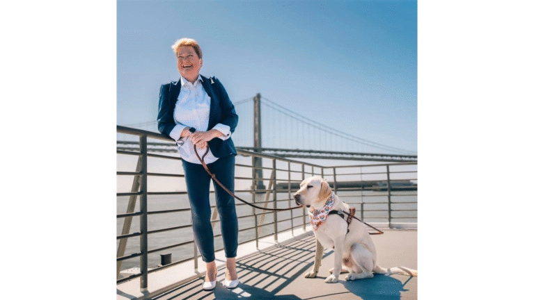 LightHouse CEO Sharon Giovinazzo stands with guide dog Pilot, a yellow lab, at the San Francisco Ferry terminal. A view of the San Francisco Bay Bridge can be seen behind her. She wears a blue pantsuit, a white-collared shirt and white heels. She holds Pilot’s leash while Pilot sits near her wearing a kerchief around the neck. The sun reflects off the top of Sharon’s short red hair as she smiles, mid-laugh.
