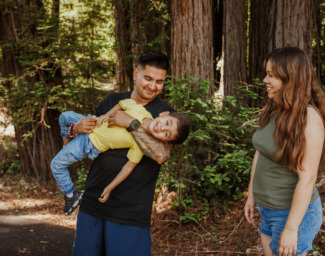 A family poses in front of the redwoods at EHC Family Camp - One parent is playing holding a laughing toddler. Photo cred: Emmalaine Berry Photography