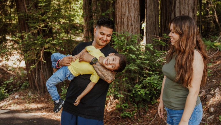 A family poses in front of the redwoods at EHC Family Camp - One parent is playing holding a laughing toddler. Photo cred: Emmalaine Berry Photography