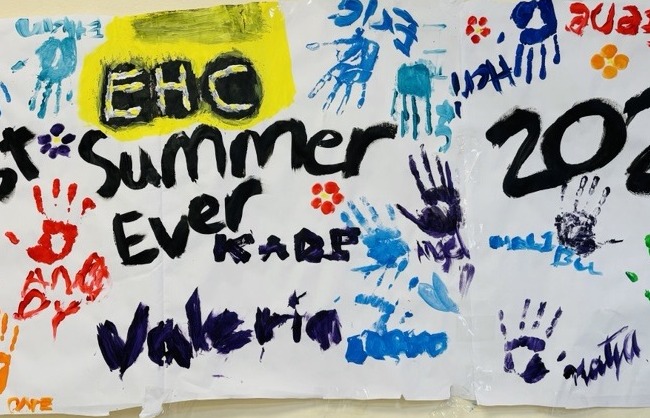 A poster on a wall at Enchanted Hills Camp features the words "Best Summer Ever 2025" and is surrounded by colorful painted hand prints, flowers, smiley faces, other doodles, and signed by the 2025 EHC Camp Staff 