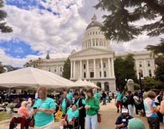 A large group of people rallying are gathered on the lawn in front of the Sacramento Capitol Building