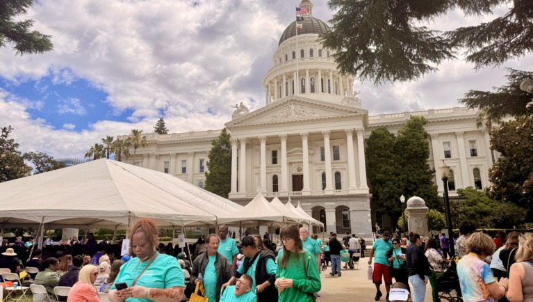 A large group of people rallying are gathered on the lawn in front of the Sacramento Capitol Building
