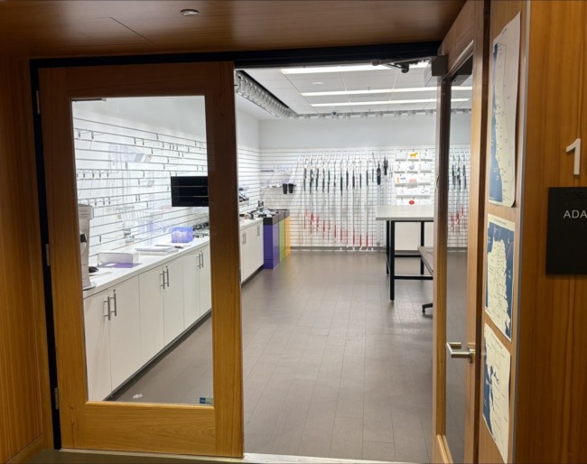 Image A storefront view into the Adaptations store, room 1090, where white canes hang on the back wall and assistive products are displayed on white slatwall panels above a long counter.