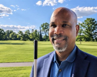 Anil Lewis, incoming LightHouse CEO, smiling outdoors in a navy blazer, holding a white cane.