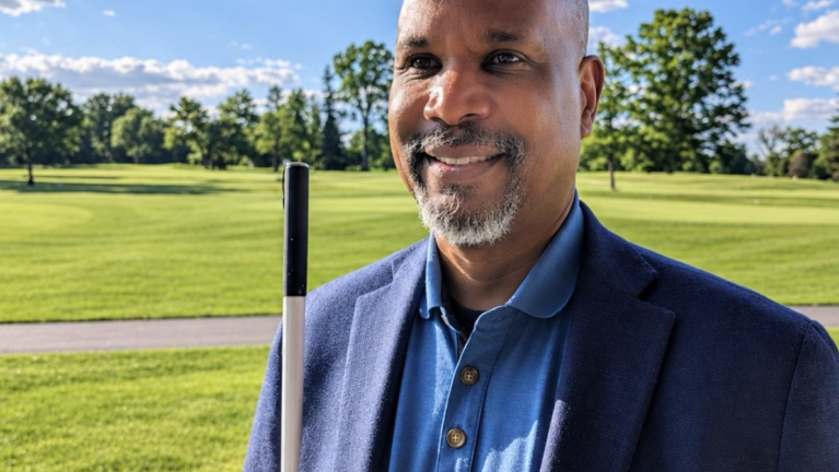 Anil Lewis, incoming LightHouse CEO, smiling outdoors in a navy blazer, holding a white cane.