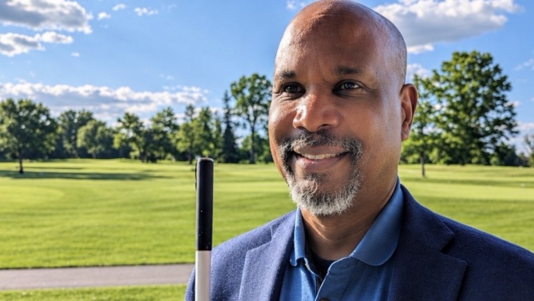 Anil Lewis, incoming LightHouse CEO, smiling outdoors in a navy blazer, holding a white cane.