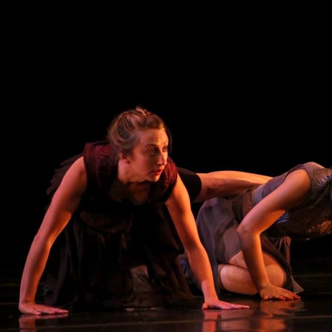 Scene from The Gift (of Impermanence): Axis Dance Company short featuring dancers with and without disabilities. Two dancers on hands and knees.  Dancer in forefront extends her leg to rest on other dancer’s back. Her expression is expectant. 