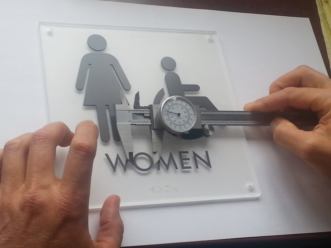 hands measure the braille on a restroom sign using calipers