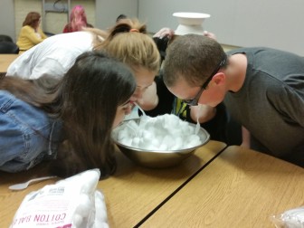 Three youth playing “cotton ball scoop” - a timed game where they had to move cotton balls, using just a spoon in their mouths, from one bowl to another bowl that is placed on a team-mates head. Each player is given 5 minutes to move as many as they can. The team with the most cotton balls at the end of 5 minutes wins.