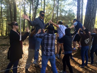 Members of the Outdoor Educators Institute (OEI) participate in a low-ropes challenge course at EHC. All under training shades, they support three members who are balancing on a single rope raised a few inches off the ground.