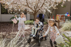 Three children smile holding Easter baskets, the child in the middle is a wheelchair user
